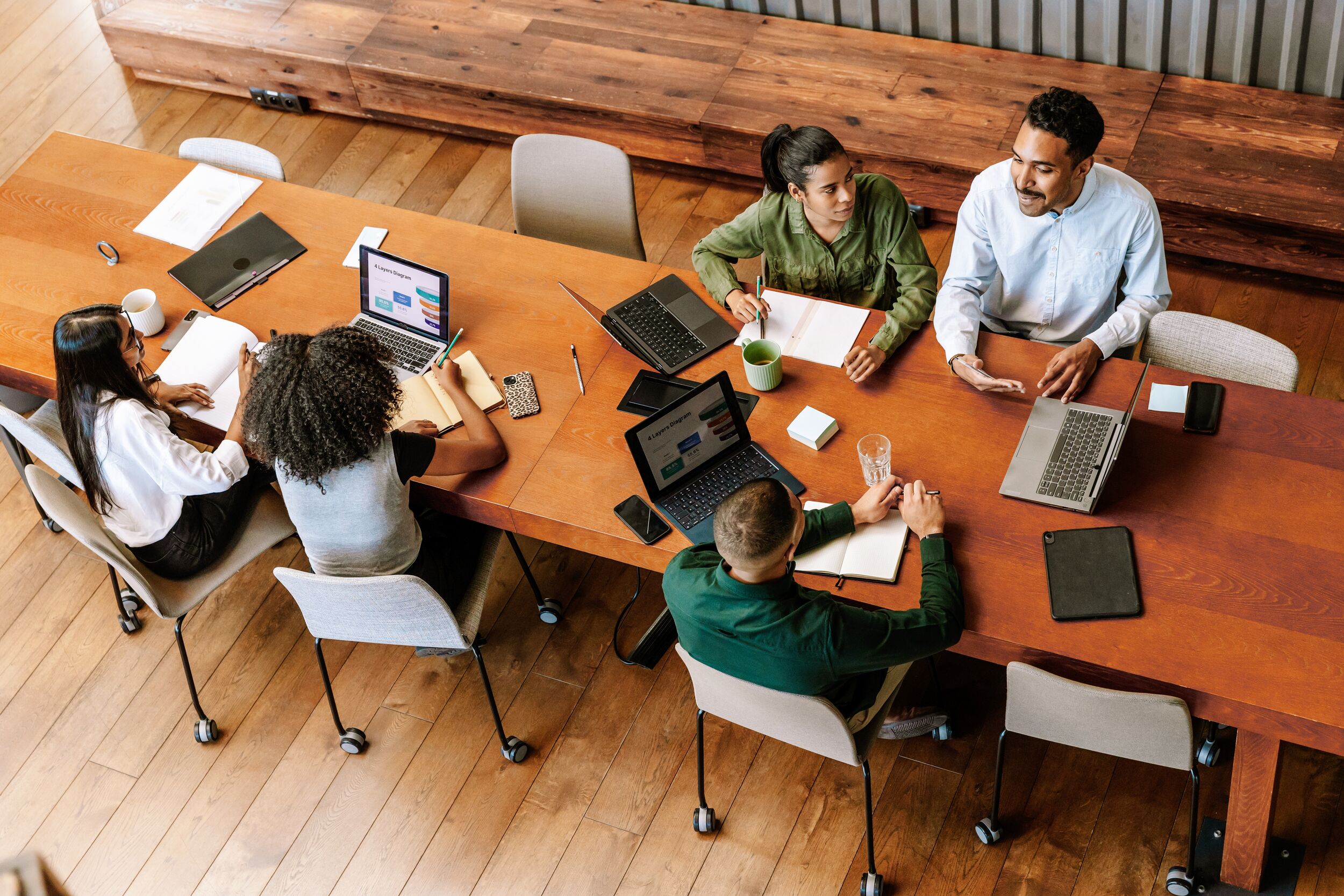 A group of three workers are talking while two female workers are looking at a laptop while sitting at a desk next to them.