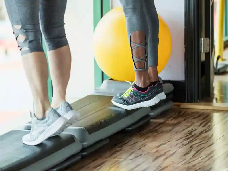 Two women standing side by side in a gym on step exercise equipment, doing calf raises.