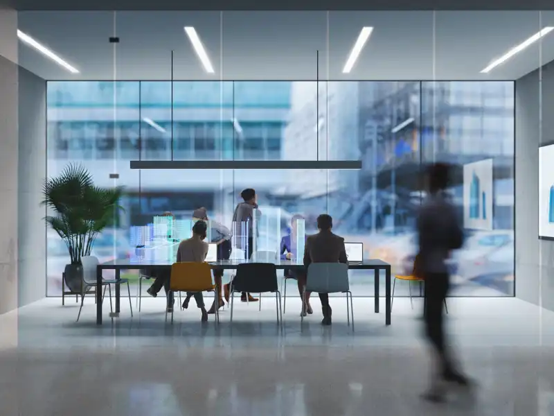 Two people sitting in a glass covered boardroom, workings while others pass by.