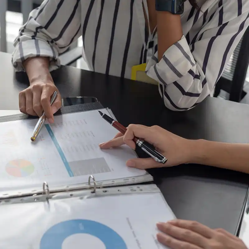 Two coworkers discuss charts and figures on a desk with an open laptop.