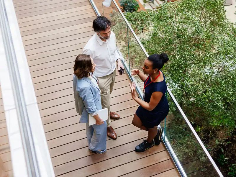 An overhead view of three people having a discussion on a wooden walkway overlooking a lush courtyard lounge area.