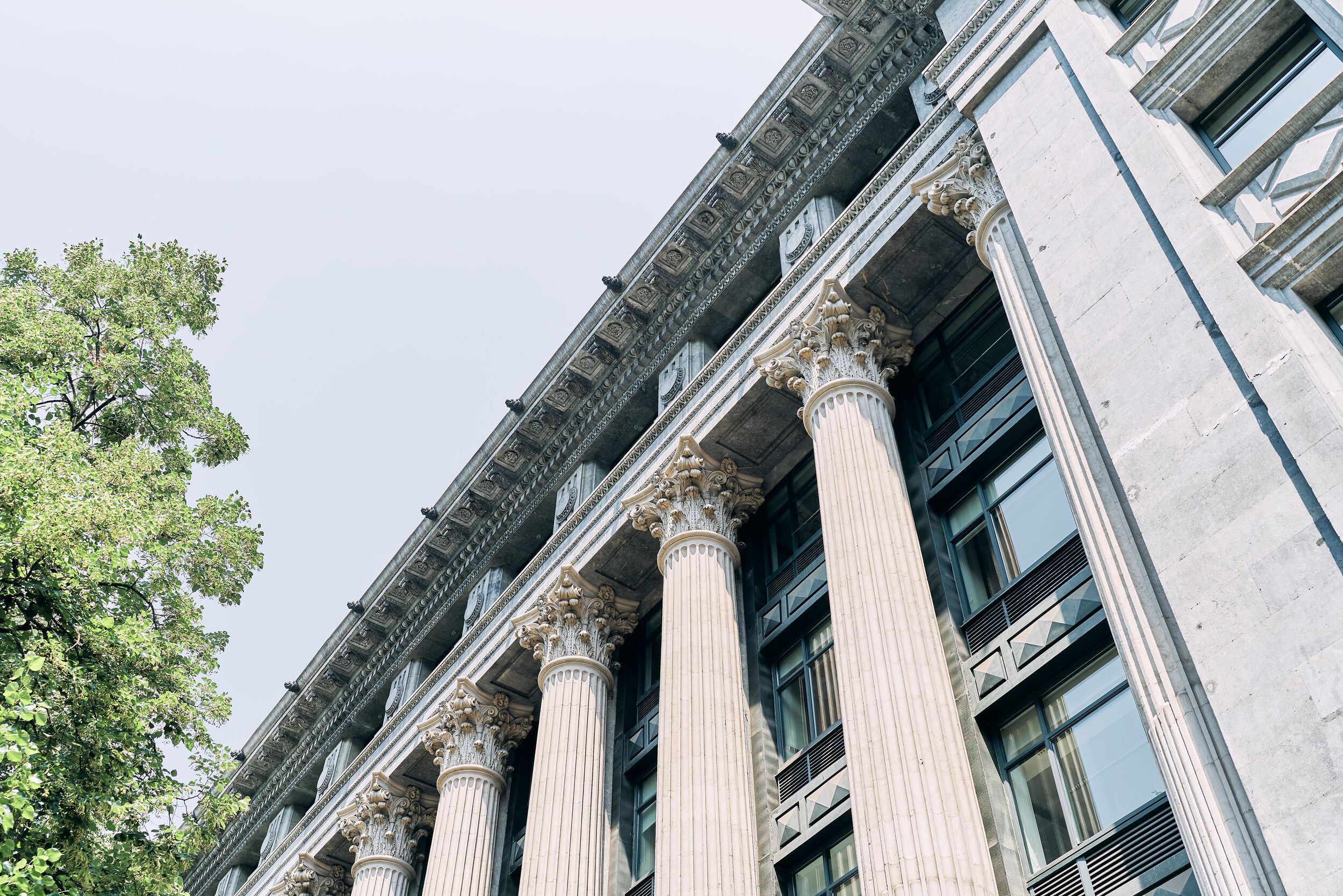 Low angle view of the exterior of a building with windows and columns with trees around, sky in background.